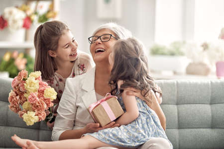 Children daughters are congratulating granny giving her flowers and gift. Grandma and girls smiling and hugging. Family holiday and togetherness.の写真素材