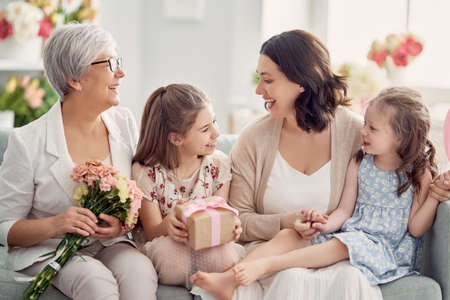 Children daughters are congratulating mom and granny giving them flowers and gift. Grandma, mum and girls smiling and hugging. Family holiday and togetherness.の写真素材