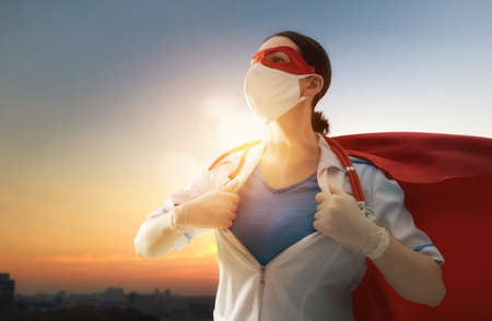 Doctor wearing facemask and superhero cape during coronavirus outbreak. Virus and illness protection, quarantine. COVID-2019. Super hero power for  medicine. Person on sunset cityscape background.の写真素材