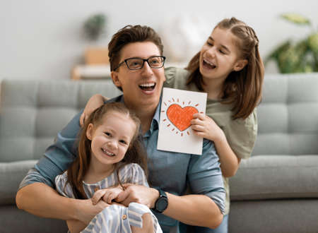 Happy father's day! Children daughters congratulating dad and giving him postcard. Daddy and girls smiling and hugging. Family holiday and togetherness.の写真素材