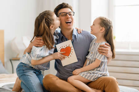 Happy father's day! Children daughters congratulating dad and giving him postcard. Daddy and girls smiling and hugging. Family holiday and togetherness.の写真素材