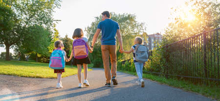 Parent and pupils of primary school are going hand in hand. Man and girls with backpacks behind backs. Beginning of lessons. First day of fall.の写真素材