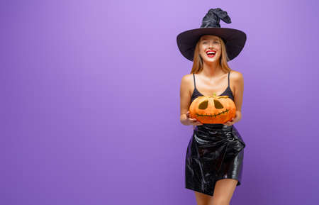 Happy Halloween! Young woman in black witch costume with pumpkin on party on purple color...