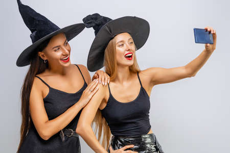Happy Halloween! Two young women in black witch costumes taking selfie on white wall background.