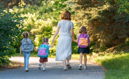 Parent and pupils of primary school go hand in hand. Woman and girls with backpacks behind backs. Beginning of lessons. First day of fall.の写真素材