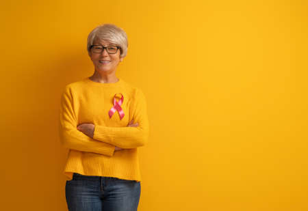 Senior woman on color yellow background. Pink ribbon like a symbol of breast cancer awareness. Support people living with tumor illness.の写真素材
