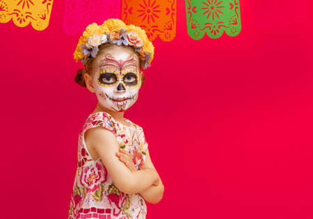 Adorable zombie in flower wreath posing on red background. Happy child with Halloween creative makeup. Girl celebrating for Mexican Day of the Dead.の写真素材