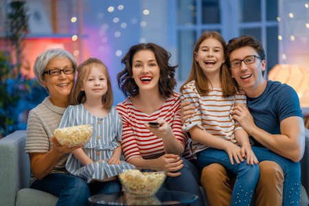 Happy family watching projector, TV, movies with popcorn in the evening at home. Granny, mother, father and daughters spending time together.の写真素材