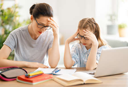 Back to school. Unhappy child and adult are sitting at desk. Girl doing homework or online...