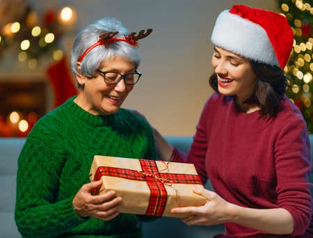 Merry Christmas and Happy Holidays! Senior mom and her adult daughter exchanging gifts. Having fun near tree indoors. Loving family with presents in room.の写真素材