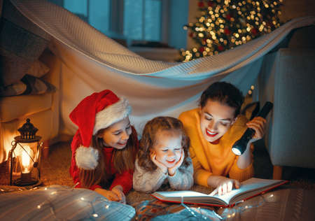 Merry Christmas! Pretty young mother reading a book to her daughters near Christmas tree.の写真素材