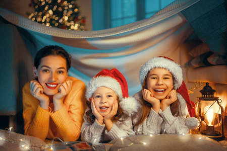 Merry Christmas and Happy Holidays! Cheerful mom and her cute daughters playing in tent. Parent and little child having fun near tree indoors. Loving family in room.の写真素材