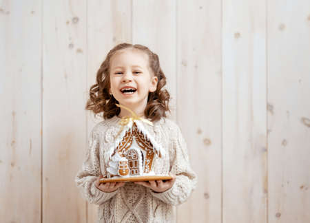 Happy little girl with Christmas gingerbread house on white wooden background.