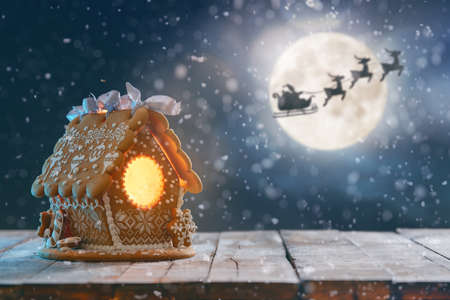 Merry Christmas and Happy Holidays! Gingerbread house on wooden table. Xmas night. Santa Claus flying in his sleigh against moon sky.の写真素材
