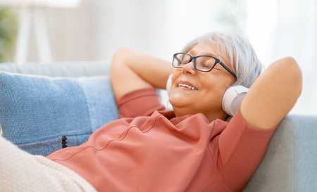 Senior woman listening to music in headphones sitting on the sofa in the room at home.