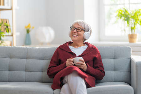 Senior woman listening to music in headphones sitting on the sofa in the room at home.