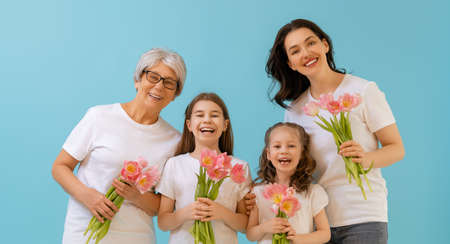 Happy women's day! Children daughters are congratulating mom and grandma giving them flowers tulips.Granny, mum and girls smiling on blue wall background. Family holiday and togetherness.