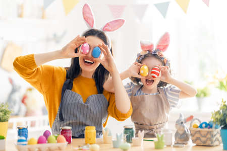 Mother and her daughter painting eggs. Happy family preparing for Easter. Cute little child girl...