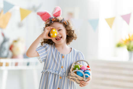 Beautiful child with painting eggs. Happy family preparing for Easter. Cute little girl is wearing...