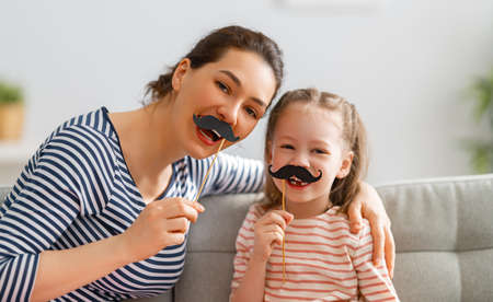 Mom and her child daughter are playing at home. Cute girl are holding paper mustache on stick. Family holiday and togetherness.の写真素材