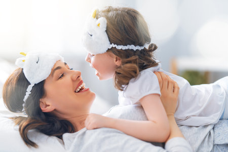 A nice girl and her mother enjoying sunny morning. Good time at home. Child wakes up from sleep. Family playing on the bed in the bedroom.の写真素材