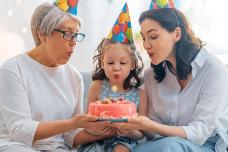 The kid is blowing out the candles on the cake. Grandmother, mother and daughter are celebrating...