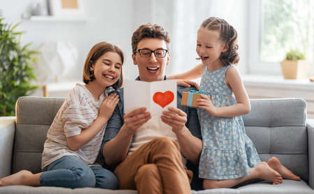 Children daughters are congratulating their father and giving him postcard. Dad and girls are smiling and hugging. Family holiday and togetherness.の写真素材