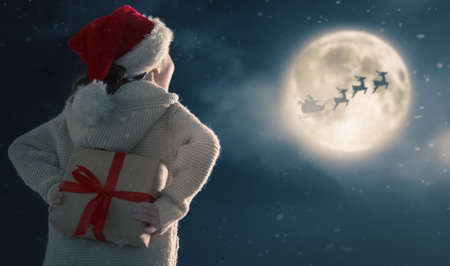 Merry Christmas. Cute little child with present. Santa Claus flying in his sleigh against moon sky. Happy kid enjoy the holiday.の写真素材