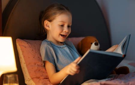 Cute little girl is reading a book in her bedroom.
