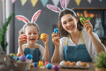 Mother and her daughter painting eggs. Happy family preparing for Easter. Cute little child girl...