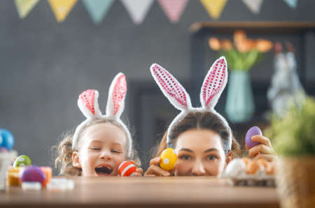 Mother and her daughter painting eggs. Happy family preparing for Easter. Cute little child girl...