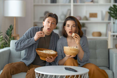 Shocked family with popcorn sitting on sofa and watching TV at home. Couple spending time together.の写真素材
