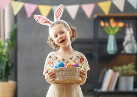 Beautiful child with painting eggs. Happy family preparing for Easter. Cute little girl is wearing...