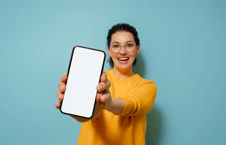 Happy young woman with smartphone on background of blue wall. Blank screen mobile phone for graphic...