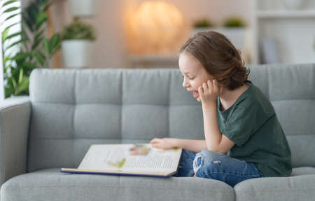 Cute little girl is reading a book sitting on the sofa.の写真素材