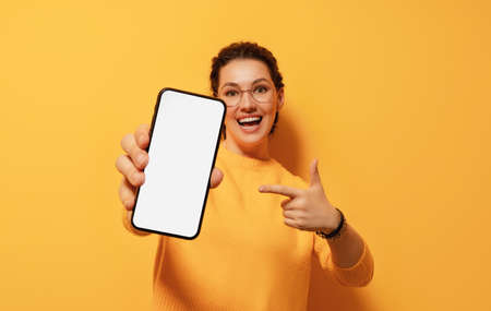 Happy young woman with smartphone on background of yellow wall. Blank screen mobile phone for...