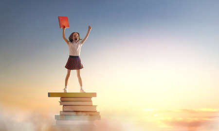 Back to school! Happy cute industrious child standing on the tower of books on background of sunset sky. Concept of education and reading. The development of the imagination.の写真素材