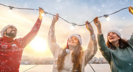 Happy friends are celebrating Christmas outdoors. Adults are having fun, hanging garlands, wearing Santa hats.の写真素材