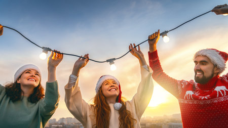 Happy friends are celebrating Christmas outdoors. Adults are having fun, hanging garlands, wearing Santa hats.の写真素材