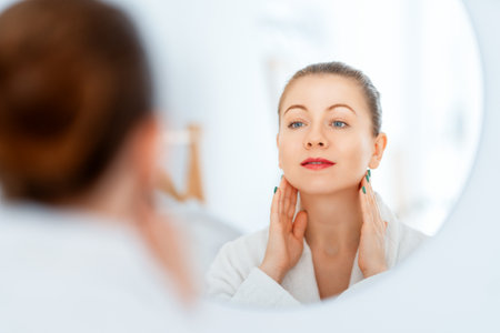 Beautiful young woman with cream for her face looking in the mirror in bathroom at home. Facial treatment. Cosmetology, beauty and spa.の写真素材