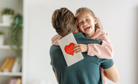 Child daughter is congratulating her father and giving him postcard. Dad and girl are smiling and hugging. Family holiday and togetherness.の写真素材