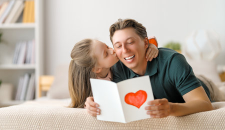 Child daughter is congratulating her father and giving him postcard. Dad and girl are smiling and hugging. Family holiday and togetherness.の写真素材