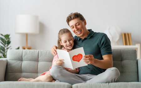 Child daughter is congratulating her father and giving him postcard. Dad and girl are smiling and hugging. Family holiday and togetherness.の写真素材