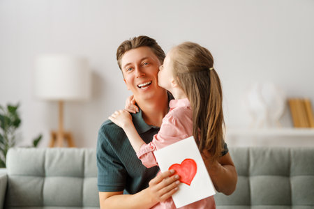 Child daughter is congratulating her father and giving him postcard. Dad and girl are smiling and hugging. Family holiday and togetherness.の写真素材