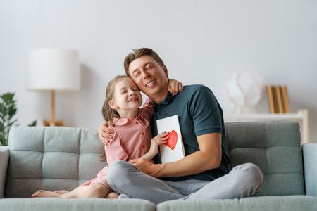 Child daughter is congratulating her father and giving him postcard. Dad and girl are smiling and hugging. Family holiday and togetherness.の写真素材