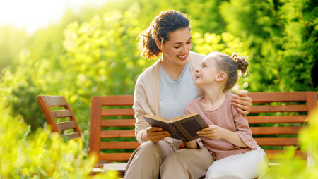 Happy little girl and her mother reading a book in summer morning. Family sitting in the garden and enjoying togetherness.の写真素材