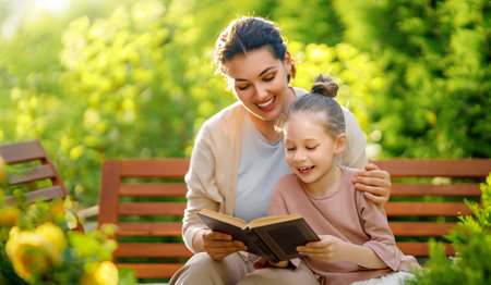 Happy little girl and her mother reading a book in summer morning. Family sitting in the garden and enjoying togetherness.の写真素材