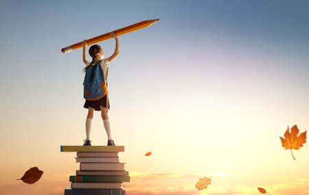 Back to school! Happy cute industrious child standing on the tower of books and holding a huge pencil on background of sunset sky. Concept of education and reading. The development of the imagination.の写真素材