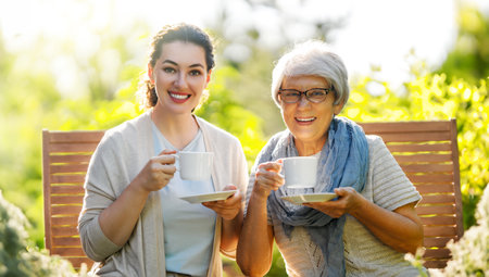 Happy young woman and her mother drinking tea in summer morning. Family sitting in the garden with cups and enjoying the conversation.の写真素材