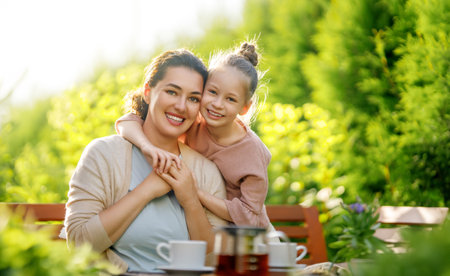 Happy little girl and her mother drinking tea in summer morning. Family sitting in the garden with cups and enjoying the conversation.の写真素材
